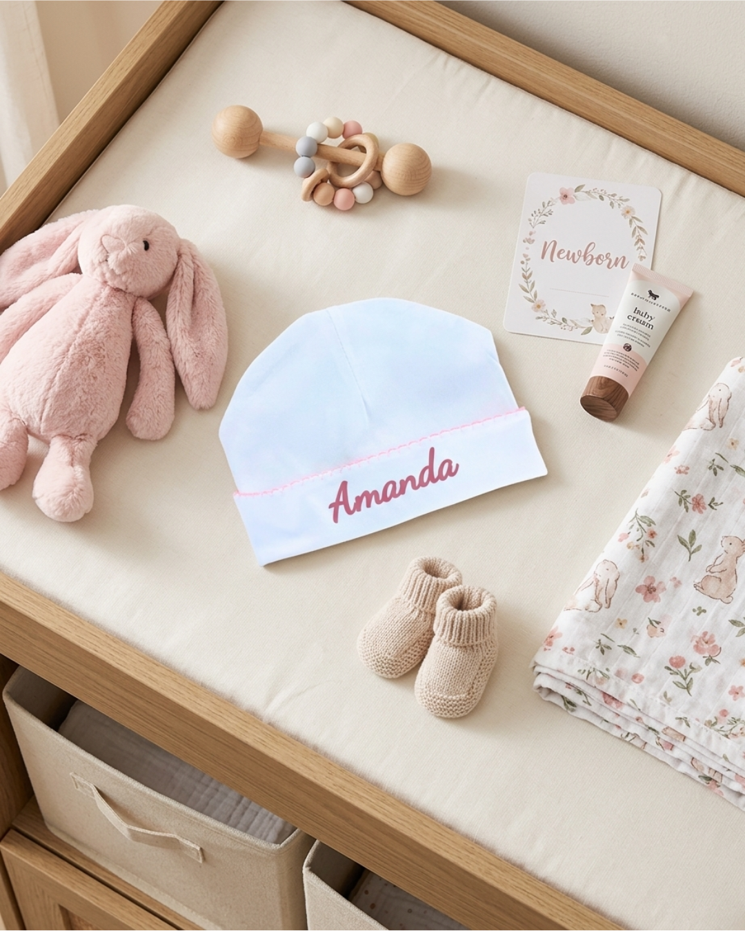 Baby items including a pink plush toy, white hat with 'Amanda' embroidered, socks, and a blanket on a wooden surface.