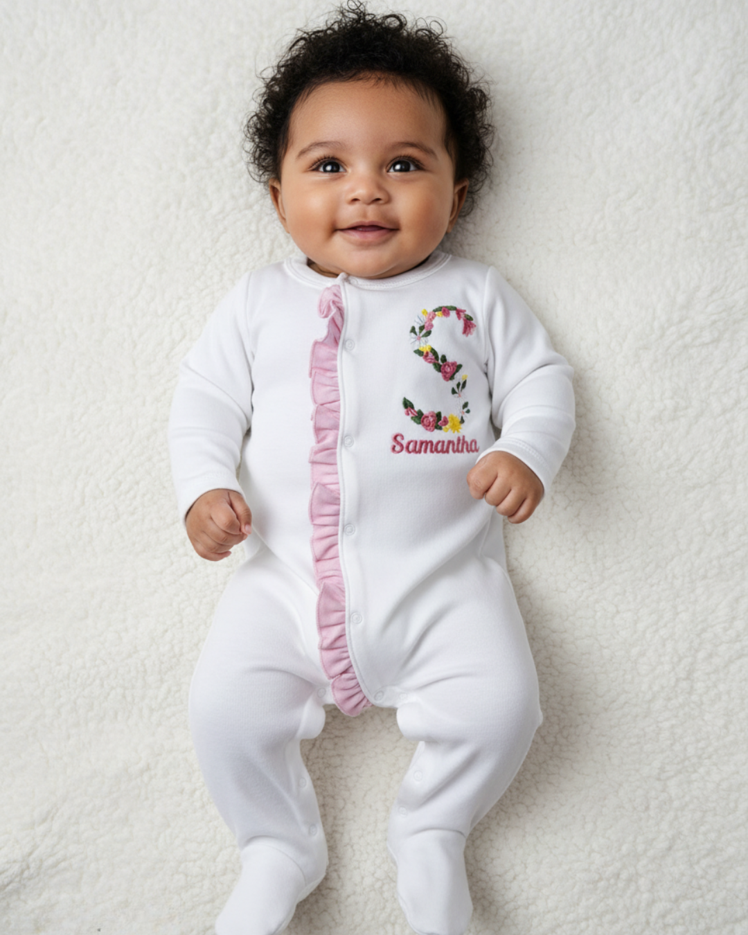 Baby wearing a white onesie with pink ruffles and floral letter 'S' design, lying on a white surface. Personalized Floral Initial Baby Girl Sleep and Play with embroidered floral letter and baby name