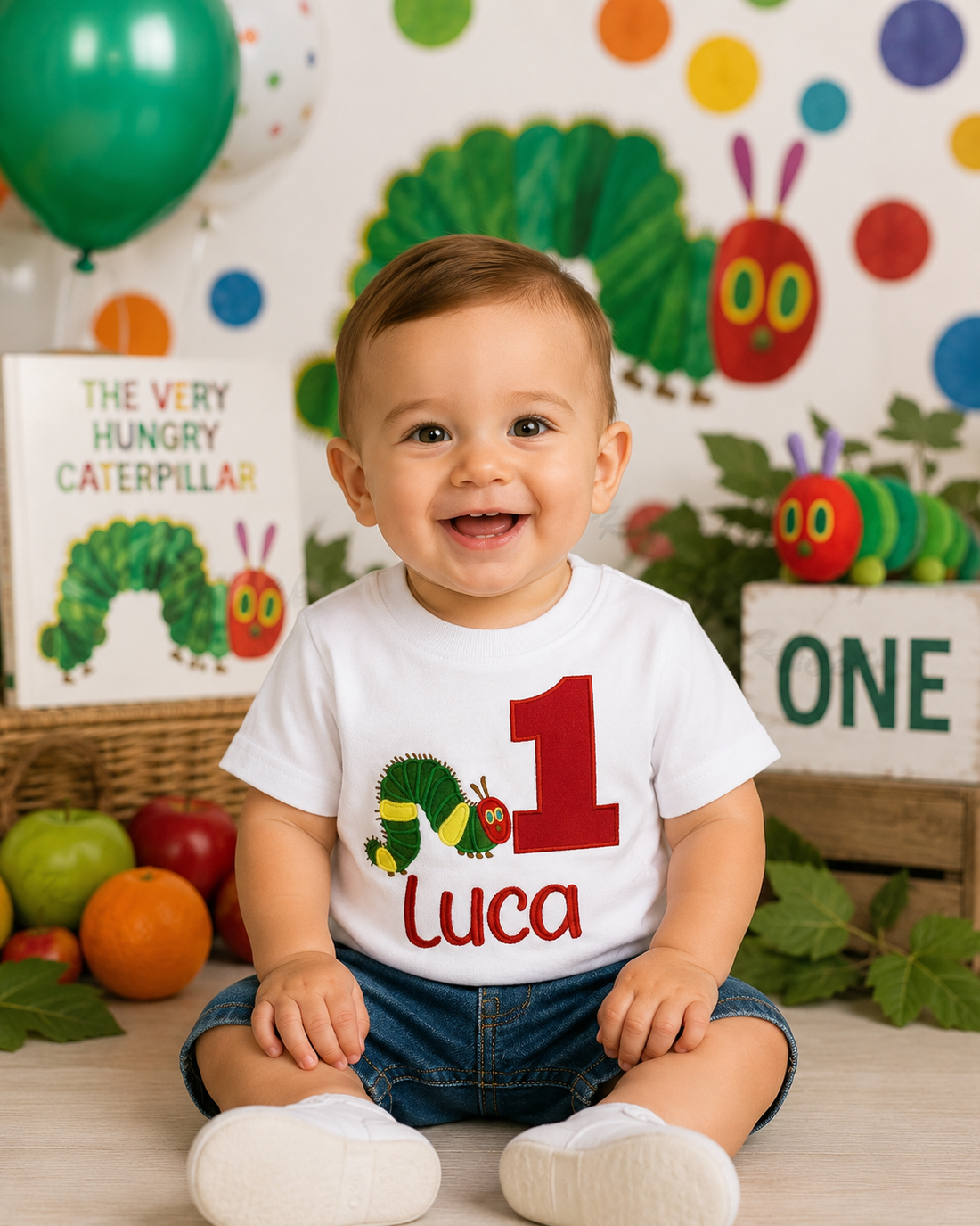 A smiling baby sitting on a floor wearing a white first birthday t-shirt with a colorful Caterpillar graphic and the name 'luca' and number '1'. The background is decorated with 'The Very Hungry Caterpillar' themed props, balloons, and a book.