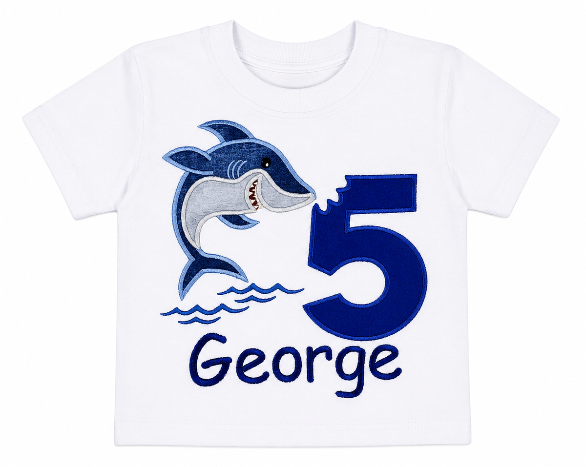 Lay-flat view of a personalized embroidered jumping shark birthday shirt featuring a blue number 5, embroidered jumping shark, and custom embroidered name George on a white 100% cotton shirt. | RoJo Custom Designs