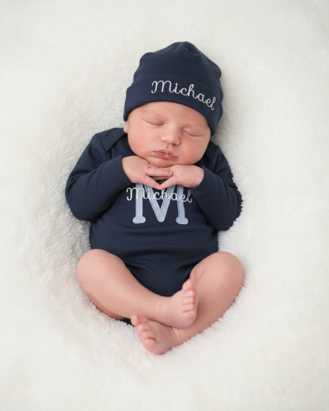 Personalized baby monogram bodysuit and hat set featuring a light blue embroidered initial and baby name on a navy cotton long sleeve bodysuit with matching embroidered hat.