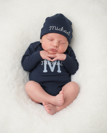 Personalized baby monogram bodysuit and hat set featuring a light blue embroidered initial and baby name on a navy cotton long sleeve bodysuit with matching embroidered hat.