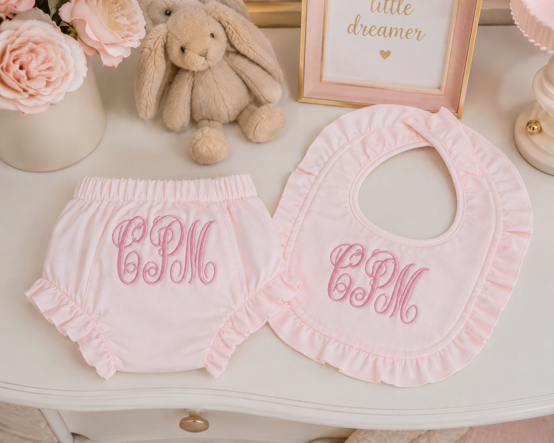 White baby diaper cover and bib set with pink embroidered monogram 'R' and floral design, styled on a cream table with a plush bunny, flowers, and framed decor.