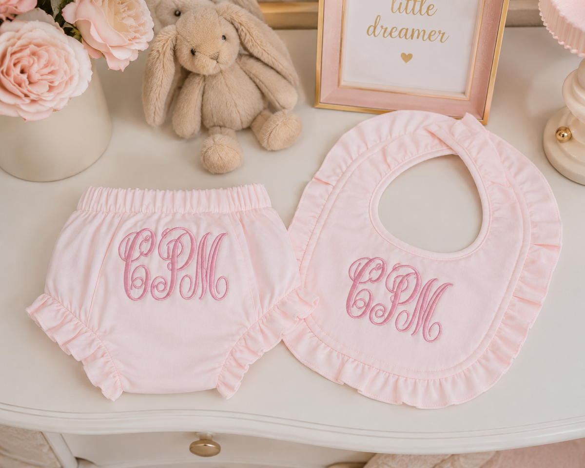 A baby outfit set featuring a light pink ruffled diaper cover and matching bib, both personalized with pink embroidered monogram letters 'CPM'. The set is styled on a white table with a plush bunny, pink roses, and a framed picture in the background.