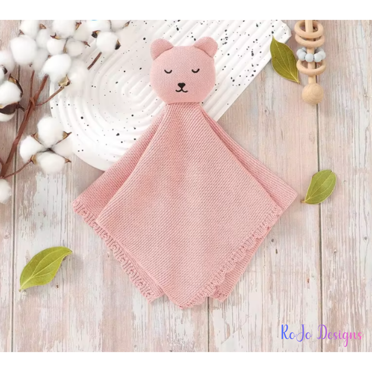 Pink bear-shaped comfort blanket on a wooden surface with cotton and leaves.