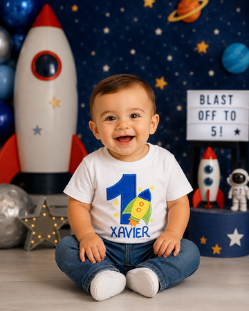 Little boy wearing a personalized embroidered rocket ship birthday shirt featuring a blue number 1, colorful rocket appliqué, stars, and custom name Xavier at a space-themed birthday party on a white 100% cotton shirt. | RoJo Custom Designs