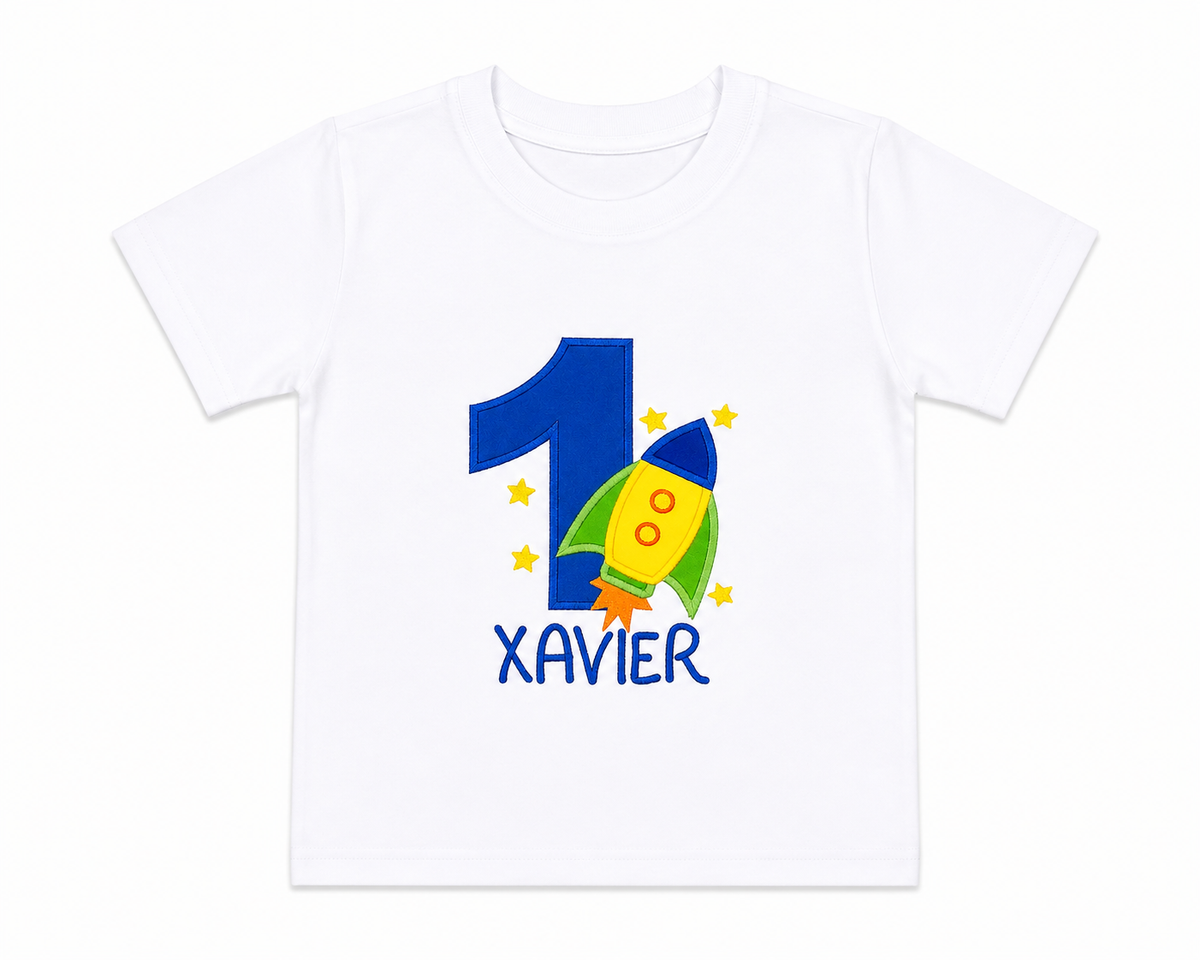 Lay-flat personalized embroidered rocket ship birthday shirt featuring a blue number 1, colorful rocket appliqué, embroidered stars, and custom name Xavier on a white 100% cotton shirt. | RoJo Custom Designs