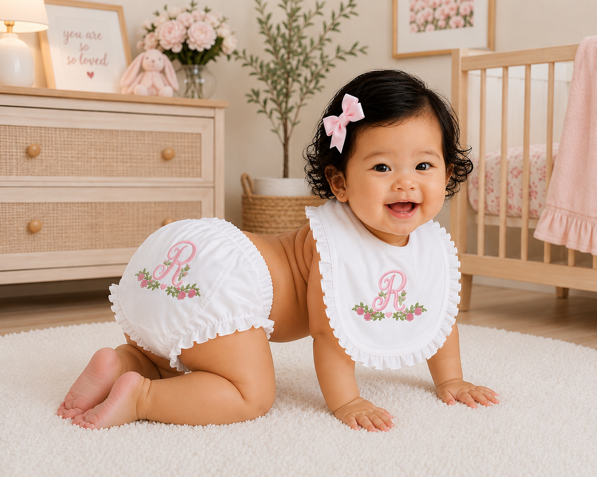 A baby crawling on a white rug in a nursery, wearing a white bib with a ruffled edge and a matching diaper cover featuring a pink monogram 'R' with floral embroidery.