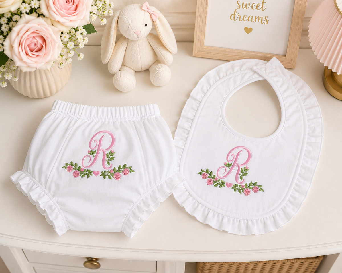 White baby diaper cover and bib set with pink embroidered monogram 'R' and floral design, styled on a cream table with a plush bunny, flowers, and framed decor.