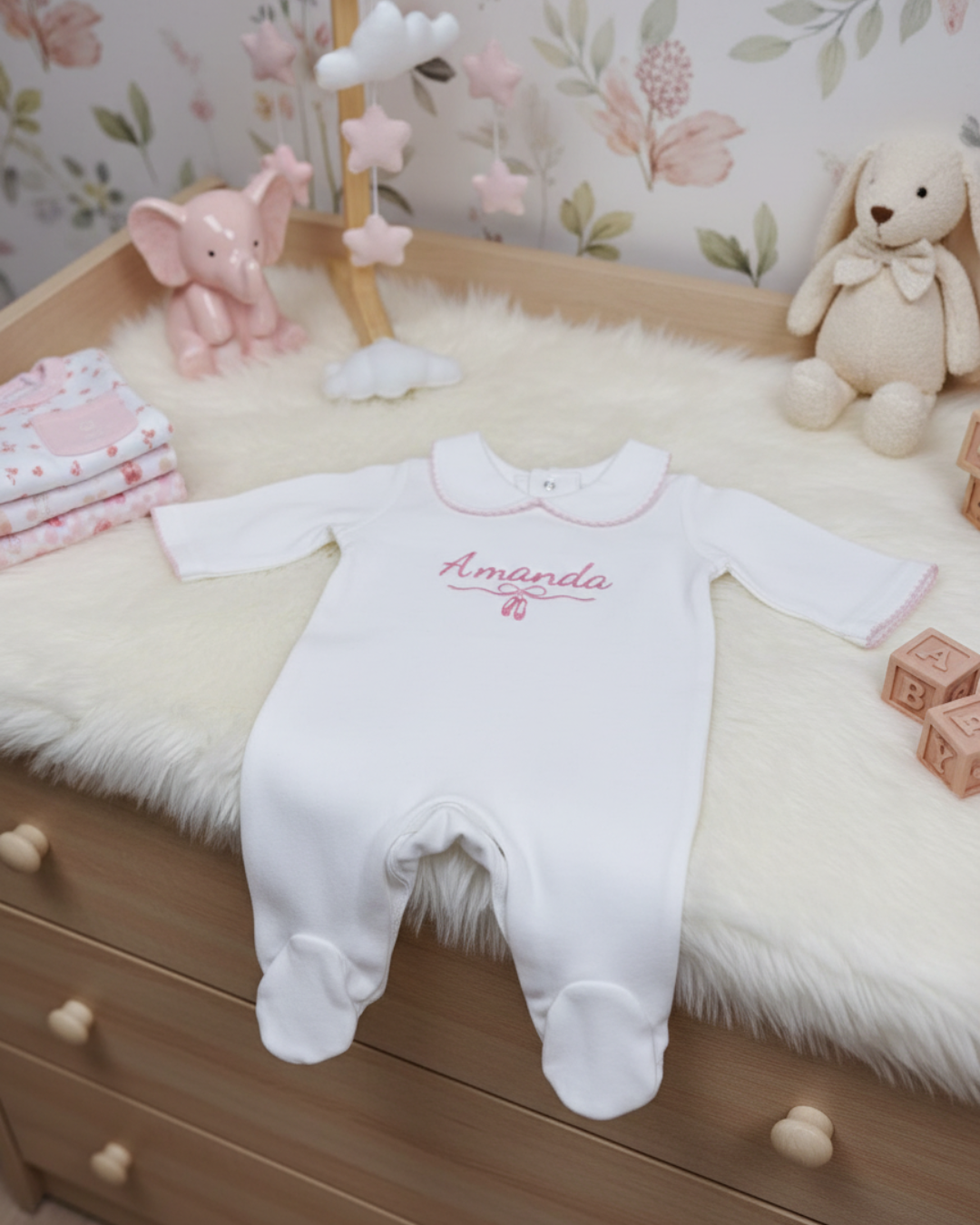 White baby onesie with 'Amelia' printed on it, placed on a wooden dresser with decorative items.