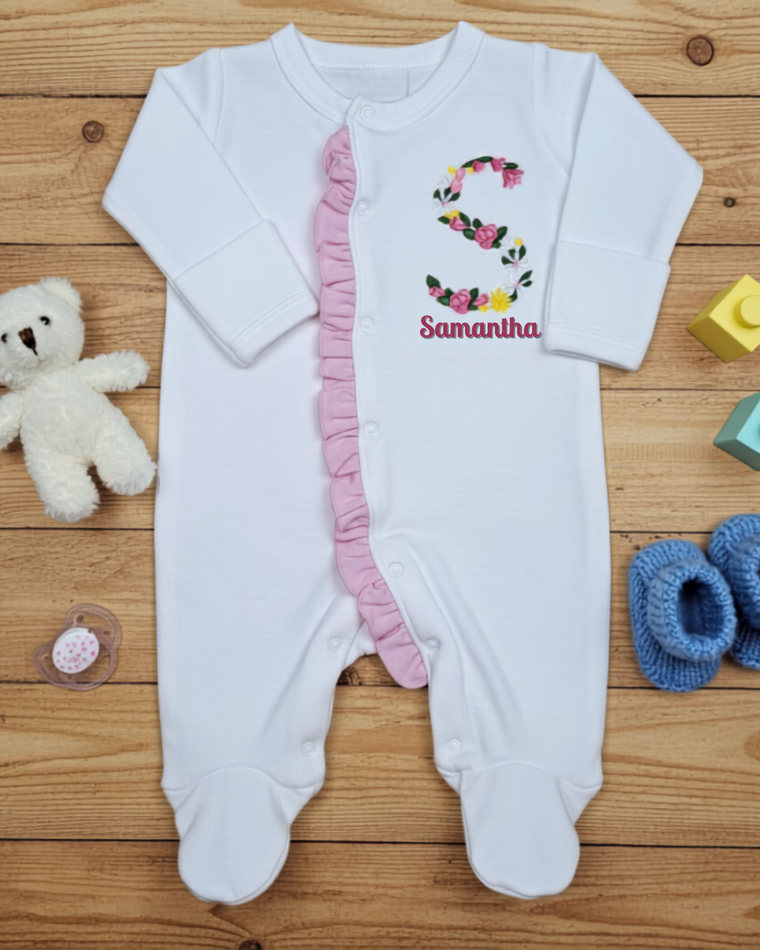 White baby onesie with floral design and name on a wooden surface with toys.